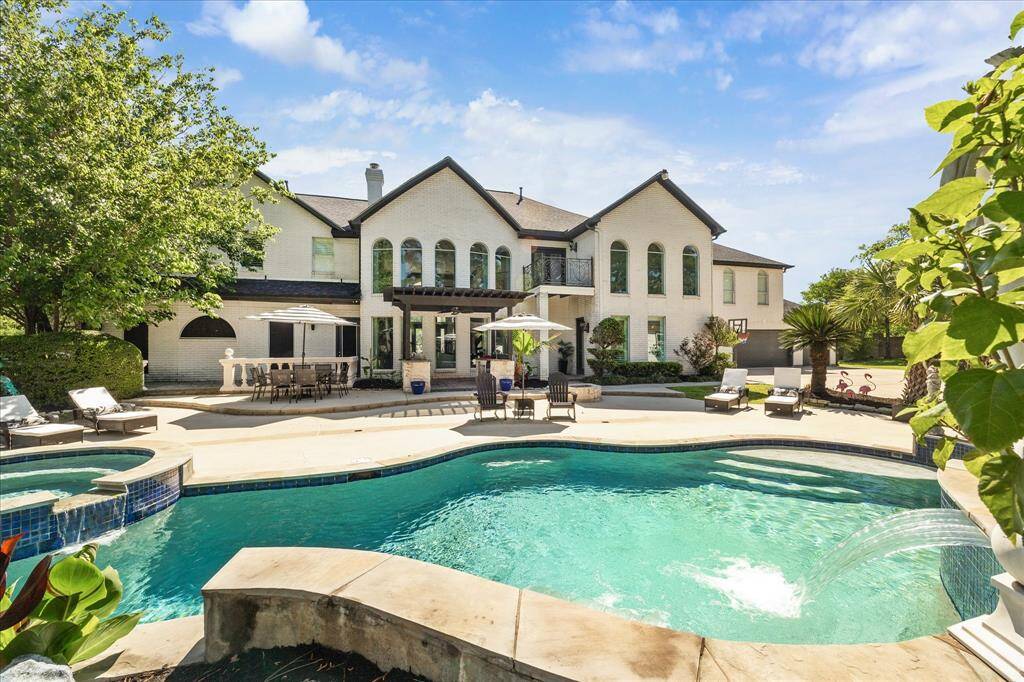 This photo showcases a luxurious backyard with a covered outdoor kitchen, featuring a built-in grill and sink. The space overlooks a sunny patio and inviting pool, surrounded by lush greenery, perfect for entertaining and relaxation.