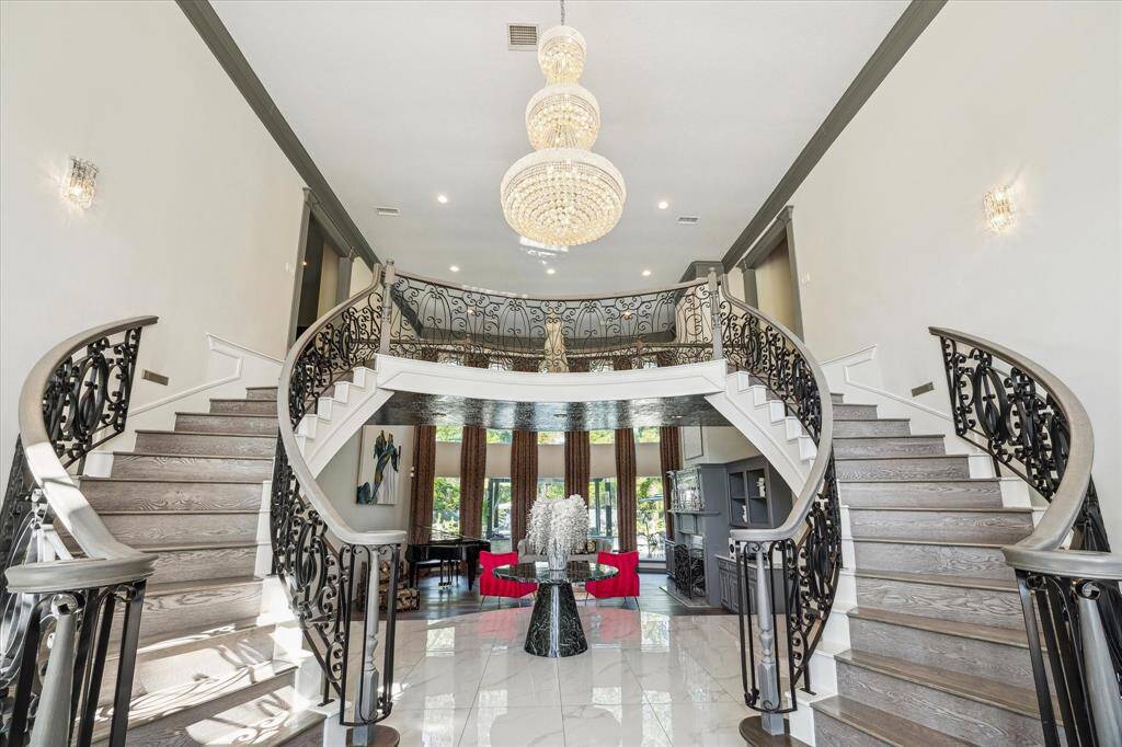 This photo showcases a grand, elegant foyer with a double staircase featuring ornate iron railings. A stunning chandelier illuminates the space, which opens into a bright living area with large windows.