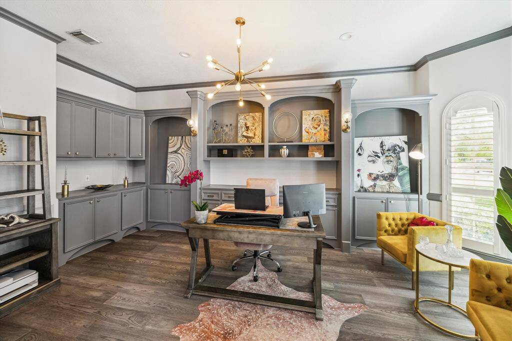 This stylish home office features elegant gray cabinetry, a modern light fixture, and ample natural light through large windows. The space is complemented by warm wood flooring, built-in shelving, and vibrant accents, creating a sophisticated and functional work environment.