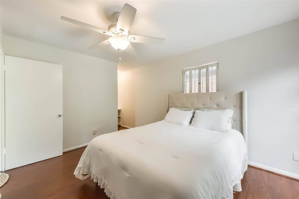 This is a cozy bedroom with a large bed, light walls, and wooden flooring. It features a ceiling fan and a small window, creating a bright and airy atmosphere.