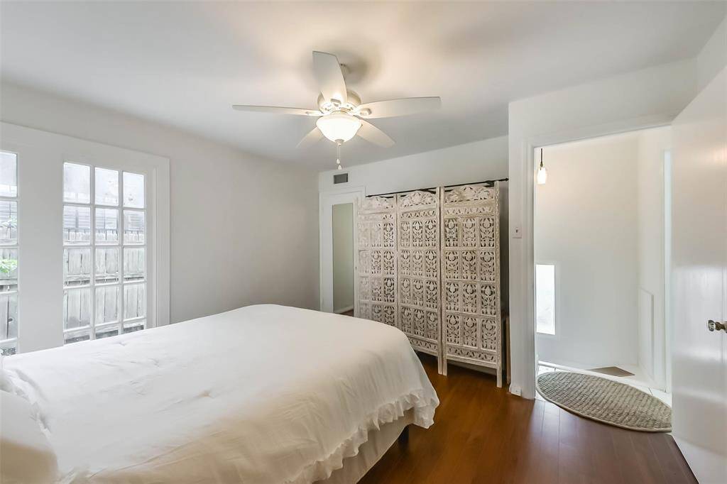 Bright, cozy bedroom with hardwood floors, a ceiling fan, and ample natural light from large windows. Features an intricately designed room divider and access to a small hallway.