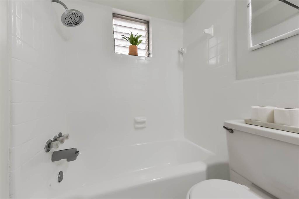 This bathroom features a clean, white design with a bathtub-shower combo. It includes a small window with natural light and a touch of greenery, adding a fresh feel to the space.