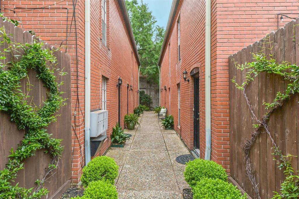 This photo showcases a charming brick pathway between two brick buildings, with neatly trimmed greenery and climbing plants on wooden fences, offering a cozy and inviting outdoor space.