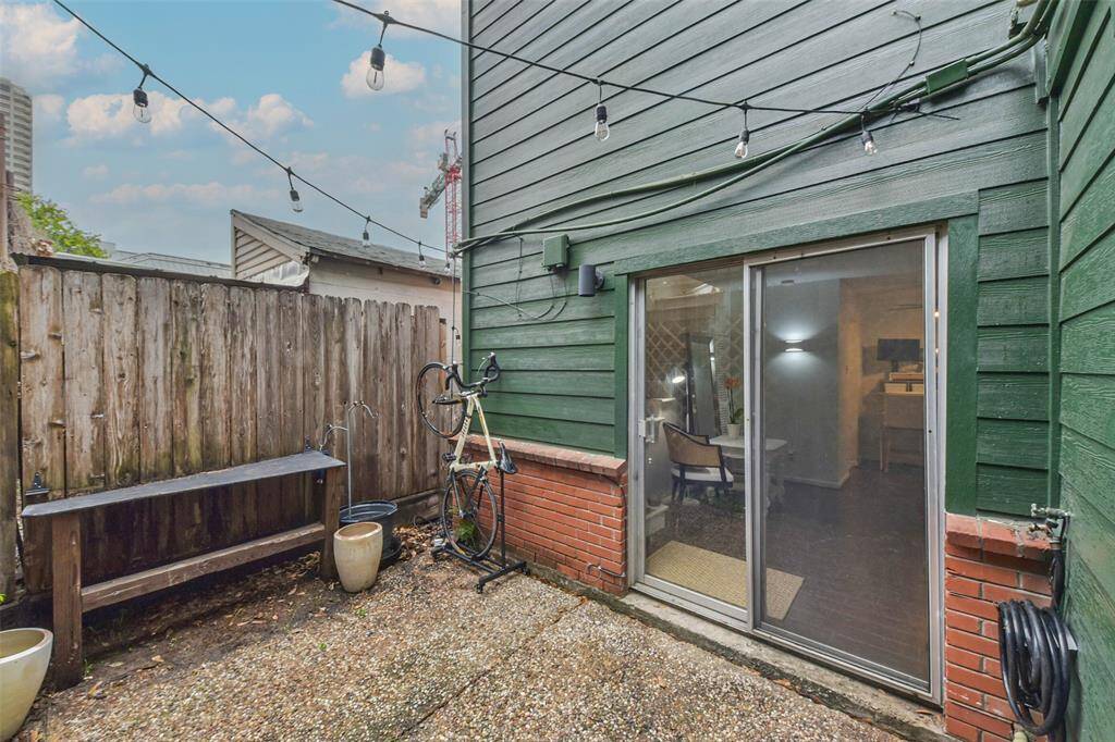 This photo showcases a cozy outdoor patio area with string lights, a wooden fence, and a sliding glass door leading inside. The space includes a small bench and planters, offering a quaint spot for relaxation or storage.