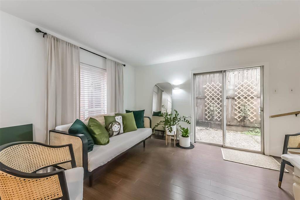 Bright, inviting living area with wood flooring, large windows with curtains, and sliding glass doors leading to a small outdoor space. Features cozy seating, green accent pillows, and decorative plants, creating a fresh, modern ambiance.
