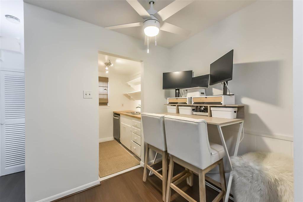 This photo showcases a small, modern workspace with two monitors on a wooden desk, accompanied by two white chairs. Adjacent is a compact kitchen with white cabinetry and a stainless steel dishwasher, illuminated by overhead lighting. The space is bright and efficient, ideal for work and cooking.