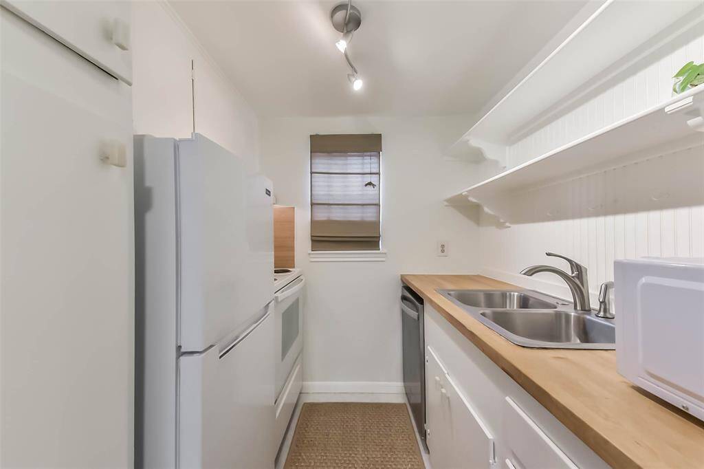 This kitchen features a compact, galley-style layout with white cabinetry and appliances. There's a wooden countertop with a double sink, open shelving for storage, and a window allowing natural light. Ideal for efficient cooking.