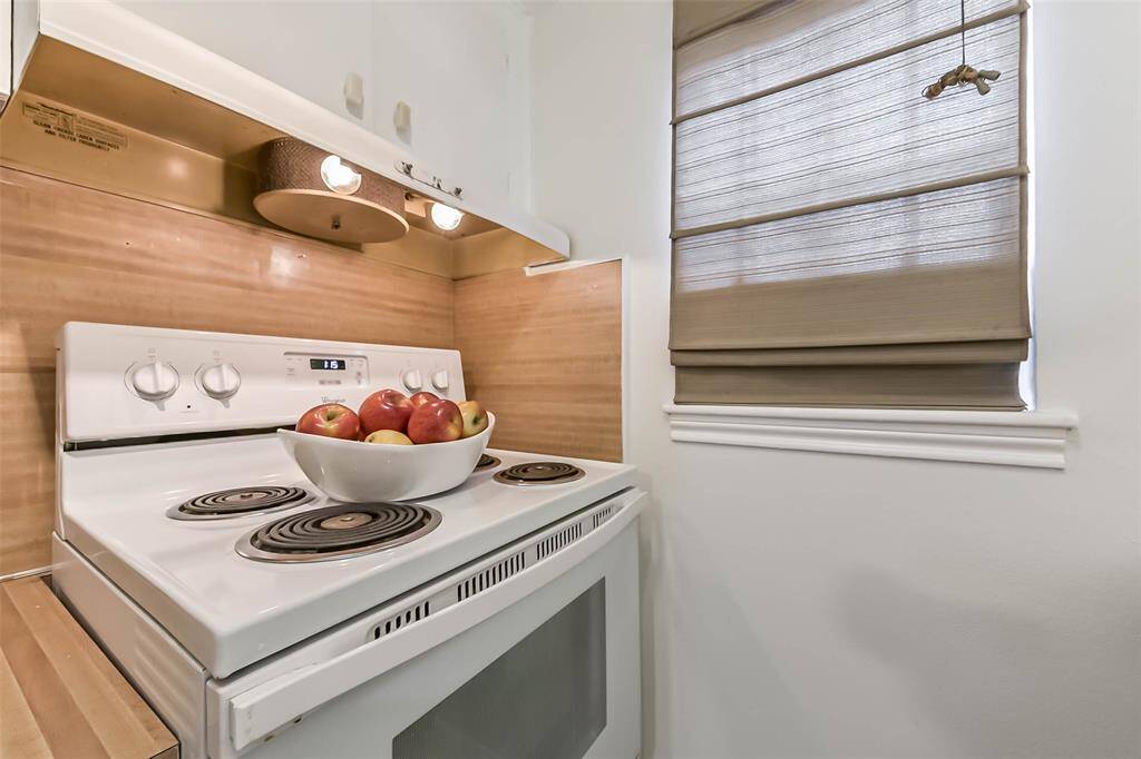 This kitchen photo features a clean, white electric stove with a bowl of apples on top, complemented by a beige backsplash and window shades, creating a cozy and inviting atmosphere.