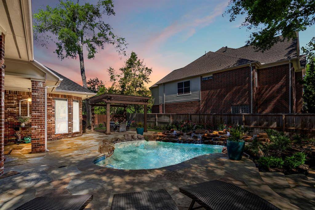 This photo showcases a charming backyard with a small pool surrounded by a stone patio. There's a cozy pergola and lush landscaping, perfect for relaxing or entertaining. The setting sun casts a warm glow, enhancing the inviting atmosphere.