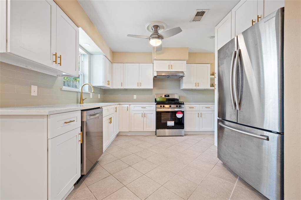 This kitchen features sleek white cabinetry, modern stainless steel appliances, quartz countertops, and a light tile backsplash. The space is brightened by large windows and includes stylish open shelving and contemporary fixtures, offering both functionality and elegance.