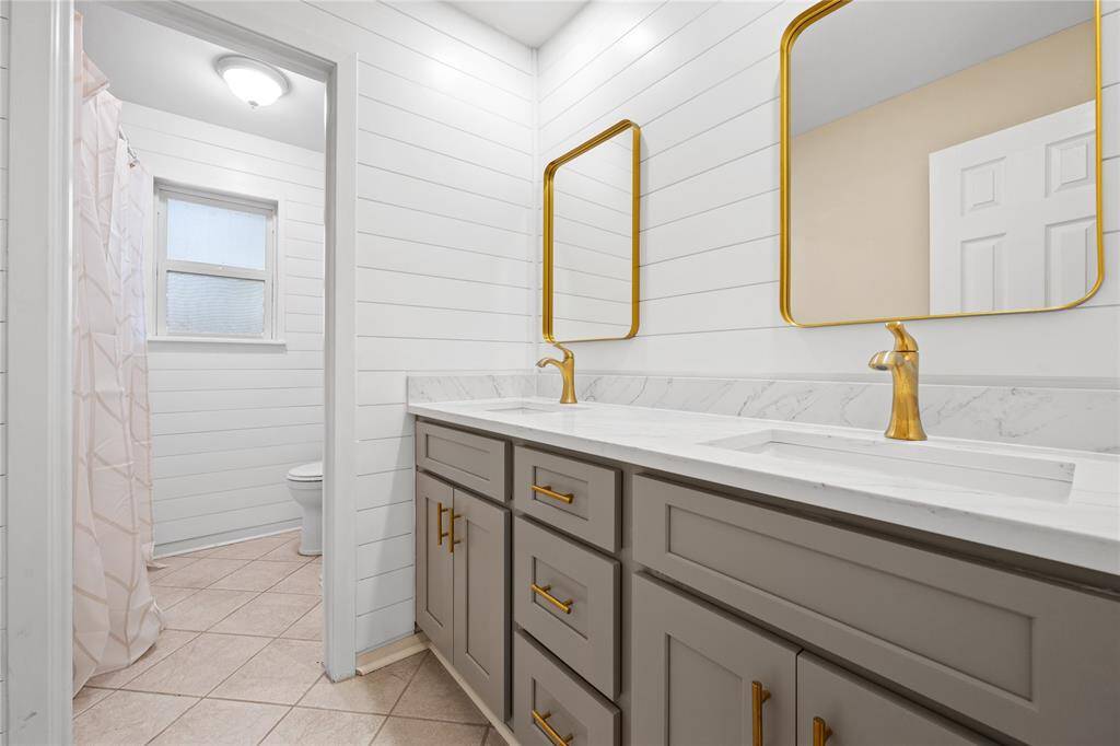 This primary bathroom features a modern design with a glass-enclosed shower, quartz countertop, stylish cabinetry with gold accents, and a large mirror above countertop. The patterned tile floor adds a touch of elegance.