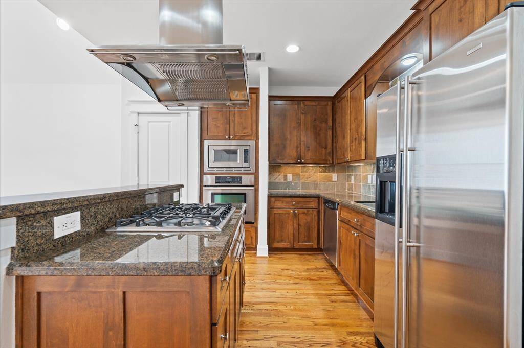 This kitchen features rich wooden cabinetry, granite countertops, and stainless steel appliances. It includes a built-in microwave, oven, and a gas cooktop with an overhead hood. The space is well-lit with recessed lighting and has a warm, inviting feel.  Refrigerator is included.