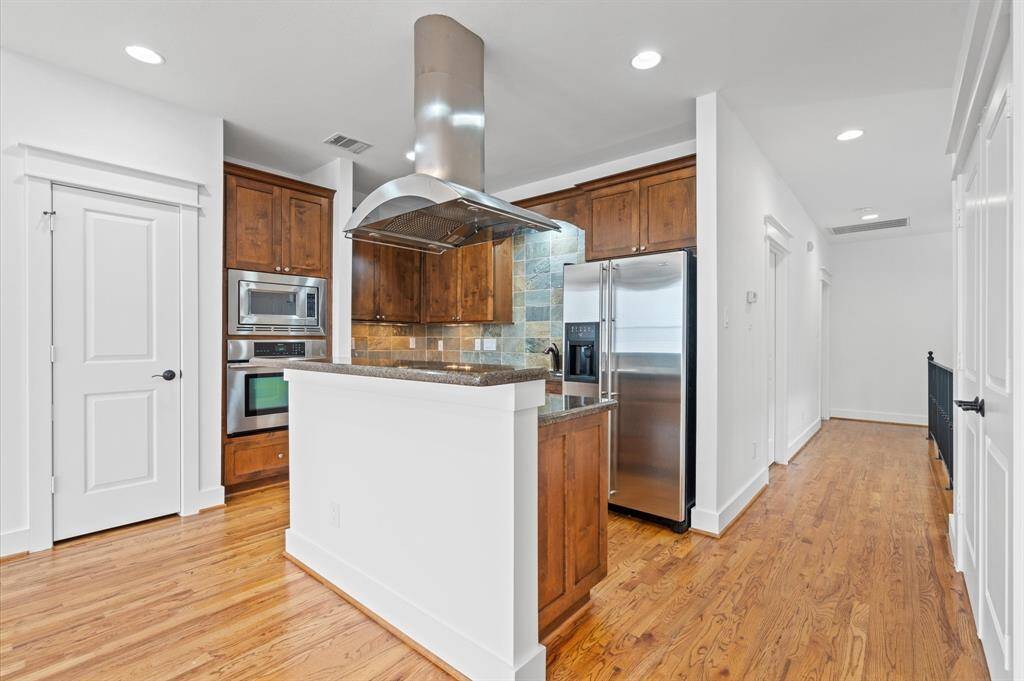 This modern kitchen features stainless steel appliances, wooden cabinets, and a sleek range hood. It has a cozy breakfast bar and hardwood floors, providing a warm and inviting atmosphere.
