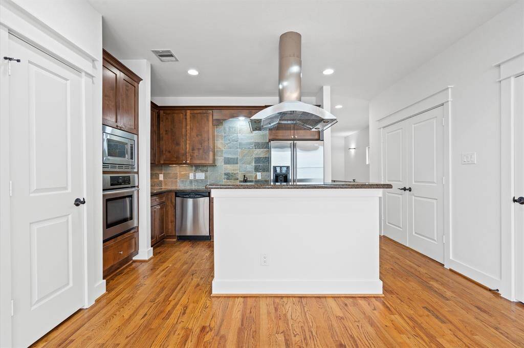 This modern kitchen features sleek stainless steel appliances, rich wood cabinetry, a central island with a stylish range hood, and a stone-tiled backsplash. The hardwood floors and ample lighting create a warm and inviting space.