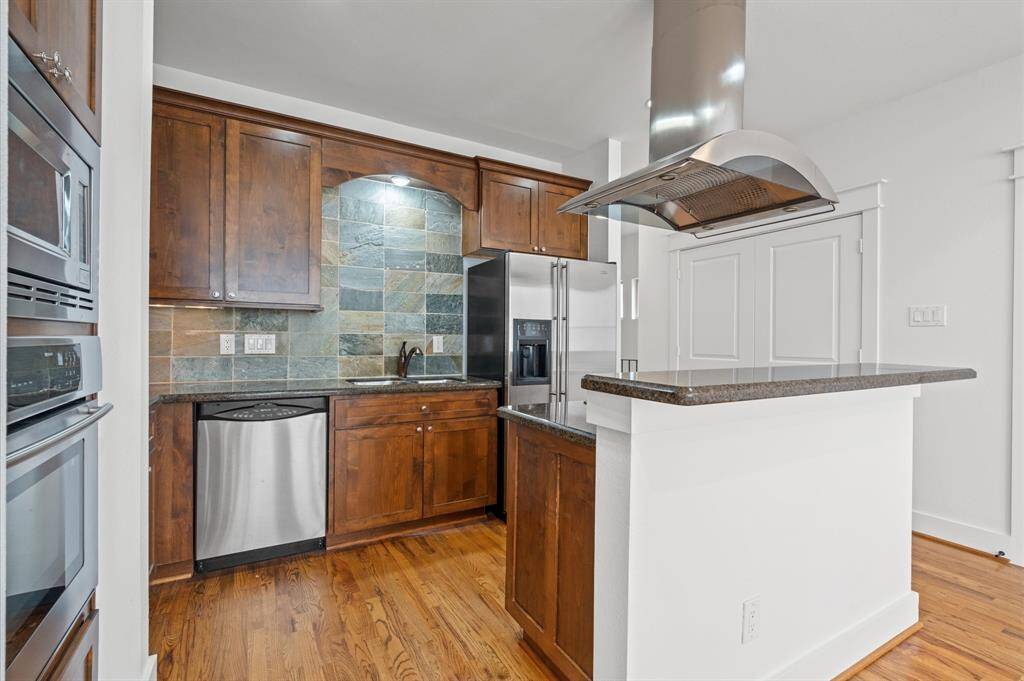 This kitchen features modern stainless steel appliances, rich wood cabinetry, and a stylish slate tile backsplash. It includes a sleek range hood over a breakfast bar, offering a blend of functionality and contemporary design. The hardwood floors add warmth and elegance to the space.