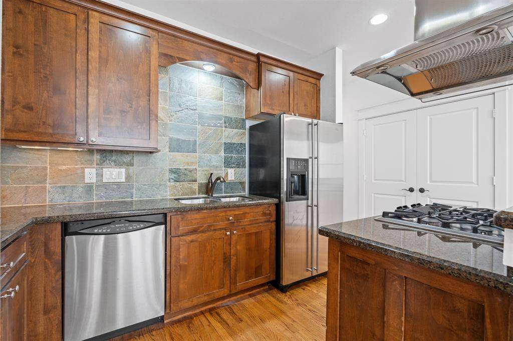 This kitchen features rich wooden cabinetry, a slate tile backsplash, and modern stainless steel appliances, including a refrigerator and dishwasher. It includes a gas cooktop with a sleek range hood and offers ample counter space on dark granite countertops.