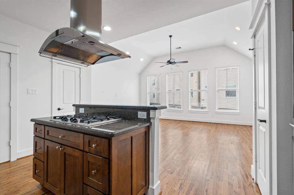 This photo showcases a bright, open-concept kitchen and living area. It features a modern cooktop island with an overhead hood, hardwood floors, and large windows with blinds, providing ample natural light. The vaulted ceiling with a ceiling fan adds to the spacious feel.