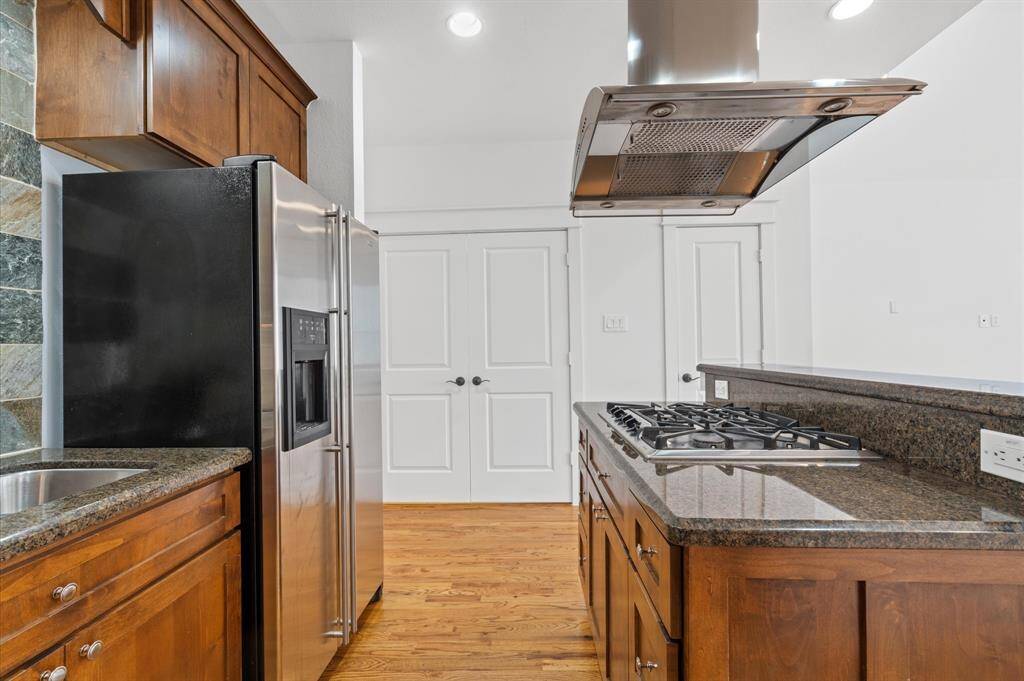 This kitchen features modern stainless steel appliances, including a fridge and a built-in gas cooktop with an overhead vent. It has warm wooden cabinetry, granite countertops, and a convenient island, all set on polished hardwood floors.