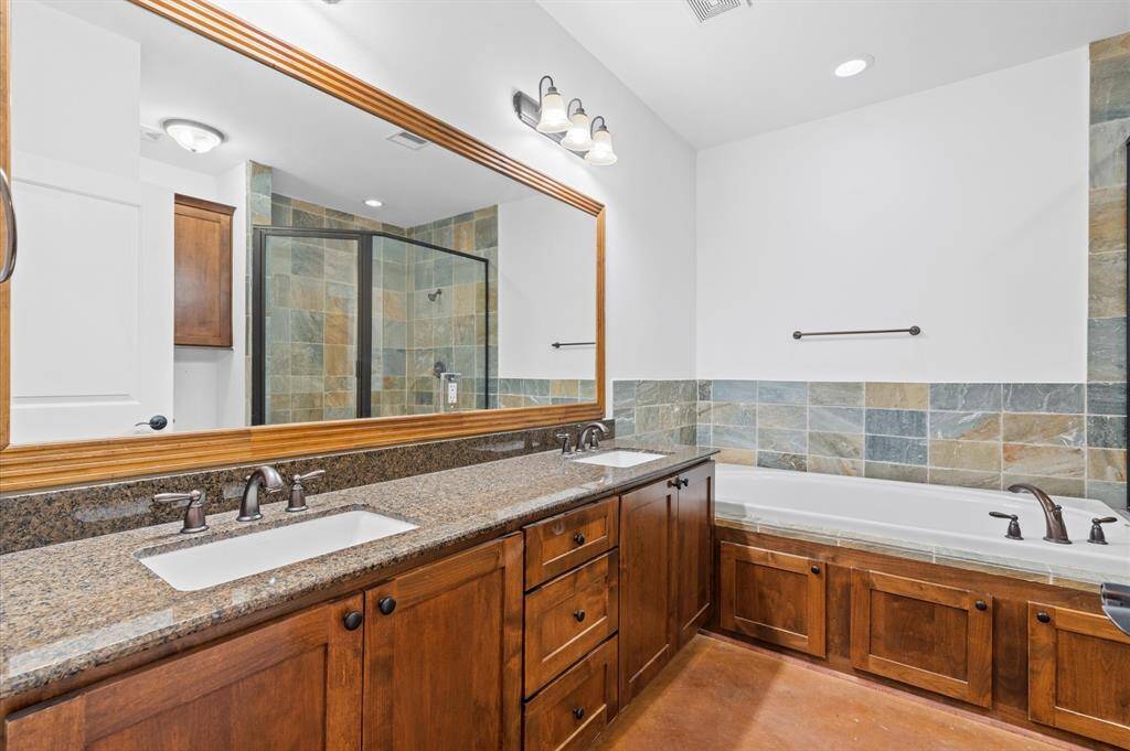 This spacious primary bathroom features a large double vanity with granite countertops and ample storage. A framed mirror enhances the space, while a separate walk-in oversized shower and a tiled bathtub provide modern comfort. The earthy tones and wood accents create a warm, inviting atmosphere.