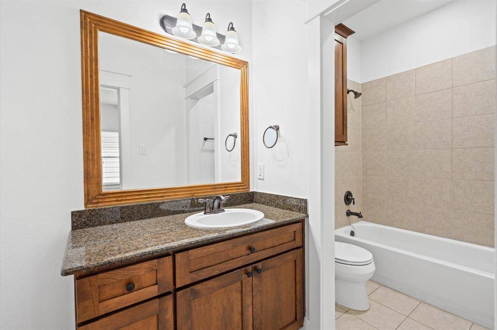 This secondary bathroom features a sleek design with a granite countertop and wooden cabinetry. It includes a large framed mirror, modern lighting, and a tiled bathtub, offering a clean and inviting space.