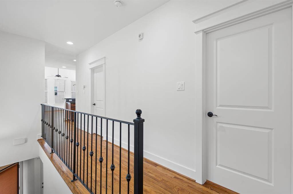 This photo shows a bright, modern hallway with white walls and hardwood floors. It features a black metal railing and leads to a room with a ceiling fan.