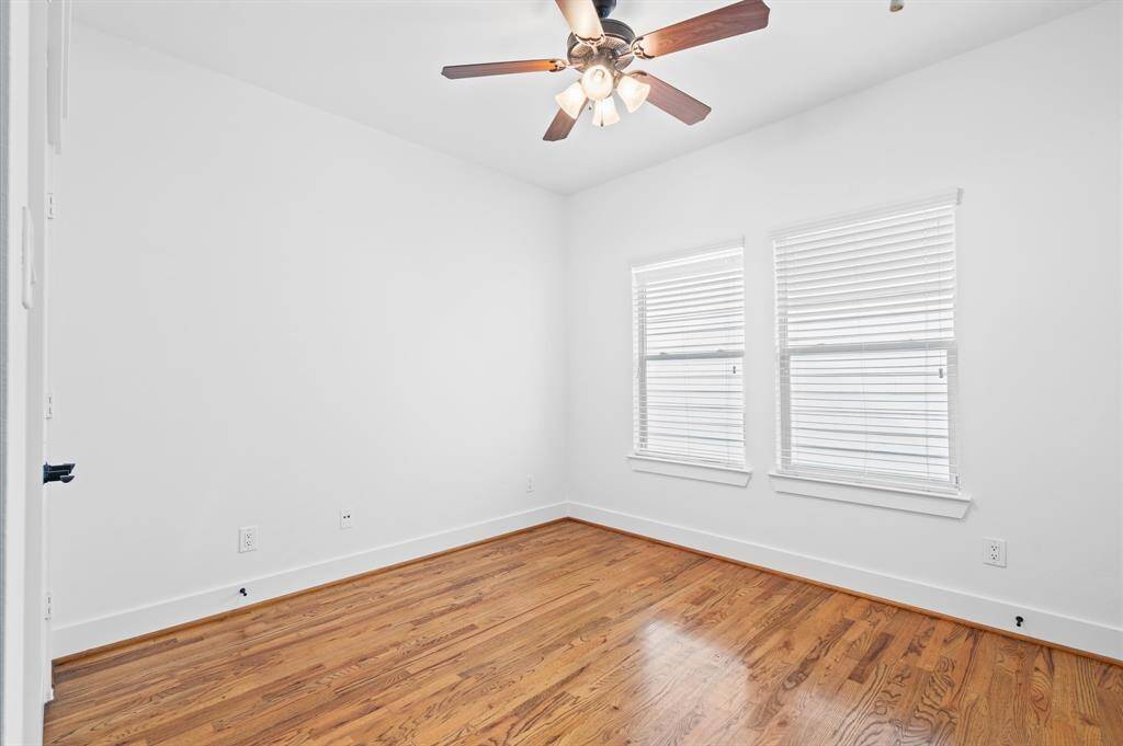 This bright secondary bedroom features hardwood floors, fresh white walls, ample natural light from two windows with blinds, and a ceiling fan for comfort. Perfect for a bedroom or home office.