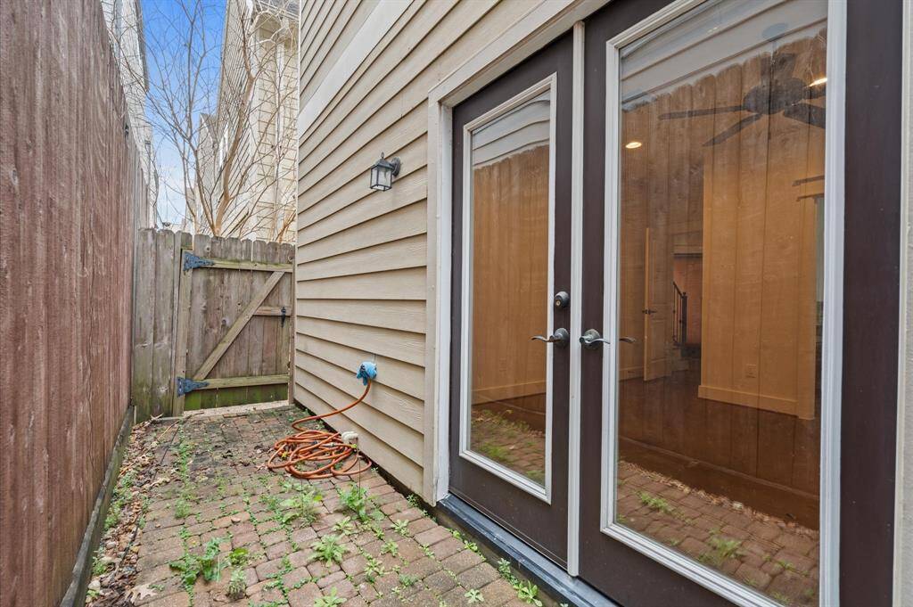 This photo shows a narrow outdoor space with brick flooring and a wooden privacy fence. It features double glass doors leading inside, a wall lamp, and a garden hose.