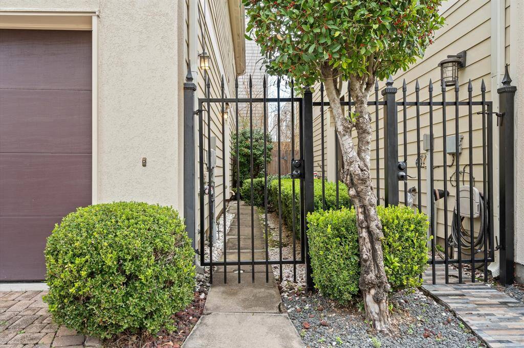 This photo shows a narrow gated side pathway between two houses, featuring a black metal gate and fence. The area is landscaped with neatly trimmed bushes and a small tree, adding a touch of greenery to the entrance.
