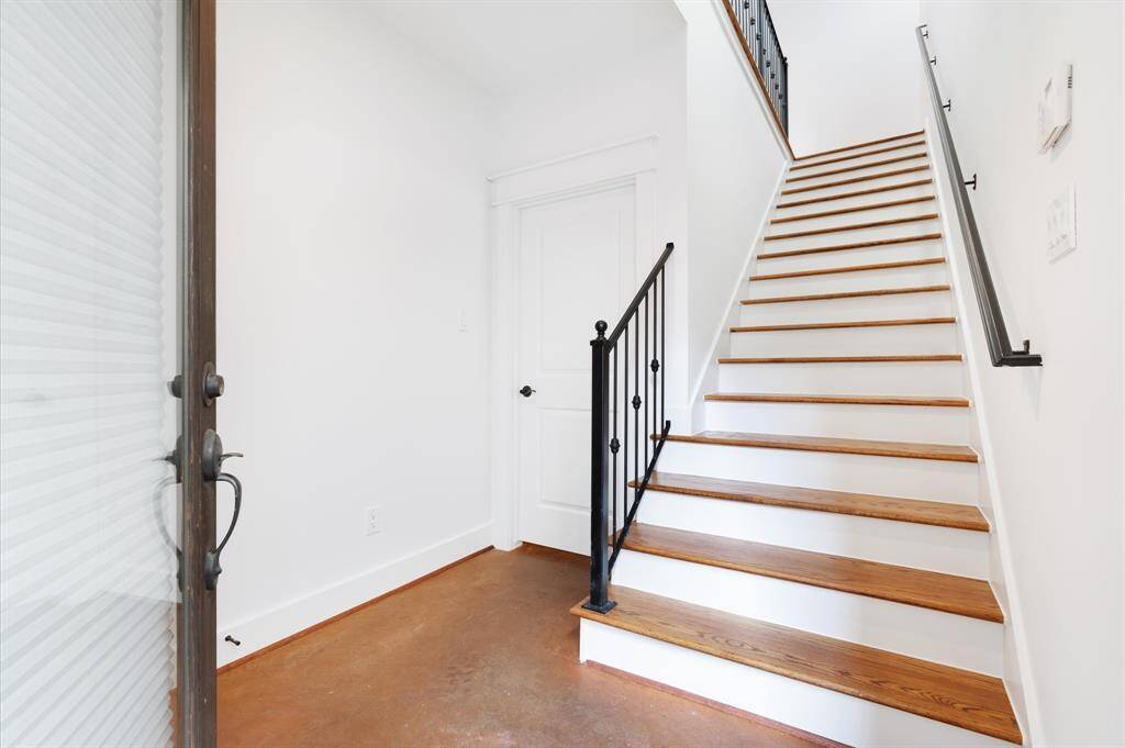 This photo shows a bright entryway with polished wooden stairs featuring a black metal railing. The space has white walls and a door, creating a clean, modern feel.