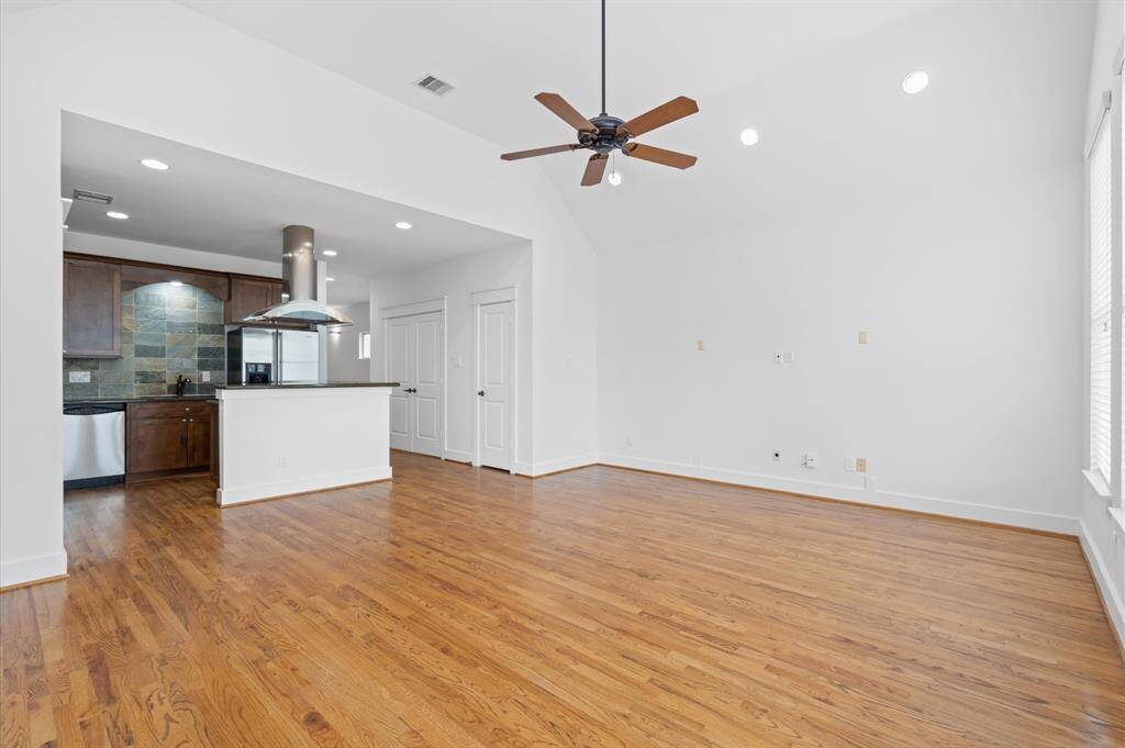 This photo showcases a spacious, open-concept living area with hardwood floors and a vaulted ceiling. The room is connected to a modern kitchen with dark wood cabinets, stainless steel appliances, and a stylish range hood. Large windows provide ample natural light, and a ceiling fan adds comfort.