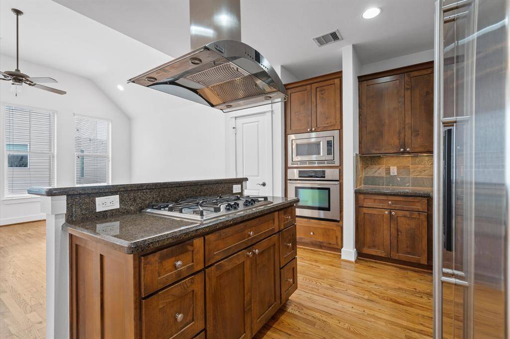 This kitchen features wooden cabinetry, a central island with a gas cooktop, stainless steel appliances, and hardwood flooring. The open layout connects to a bright family area with large windows and a ceiling fan.