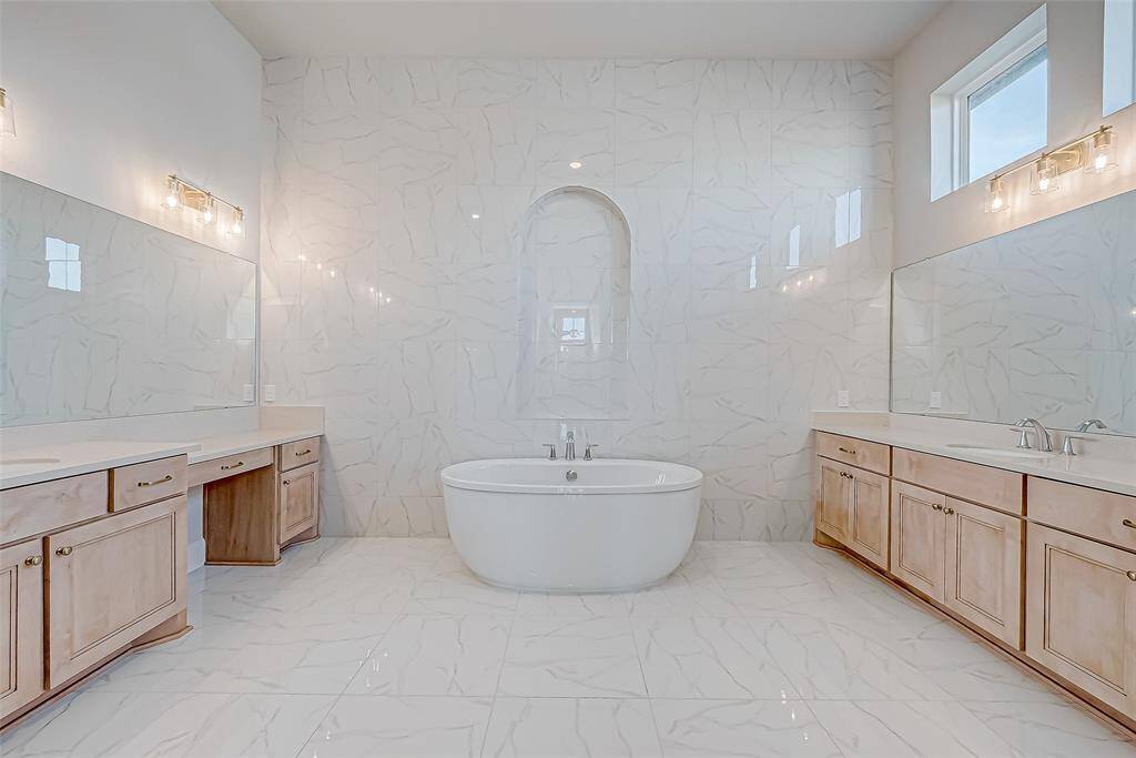 The ensuite bathroom is a spa-like retreat with a freestanding tub framed by a fully tiled wall w/ a niche, a fully tiled super shower with a rainfall system, and double vanities with natural wood cabinetry and expansive mirrors. Two immense walk-in closets flank the hallway leading to the bedroom, offering exceptional storage.
