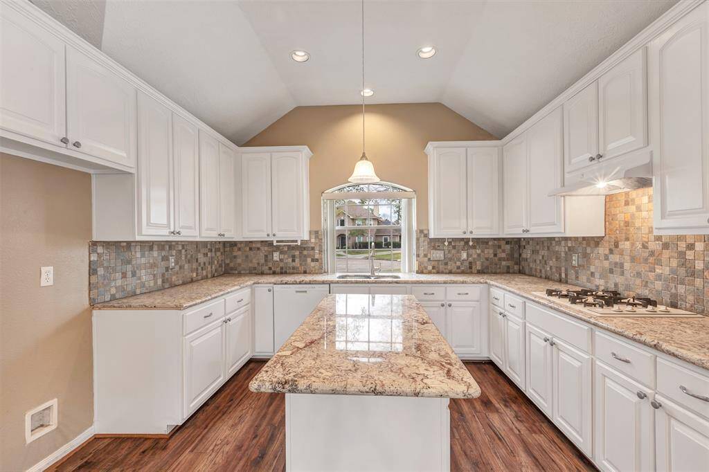 This kitchen features elegant white cabinetry, granite countertops, and a stylish tile backsplash. The space is well-lit with recessed and pendant lighting, complemented by a central island and gas stove. The warm wood flooring adds a cozy touch.