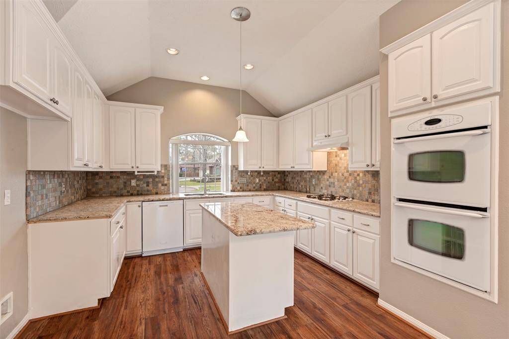 Bright, spacious kitchen with high ceilings, featuring white cabinetry, granite countertops, and a central island. Equipped with modern appliances, including double ovens, and a large window providing natural light. Warm wood flooring adds a cozy touch.