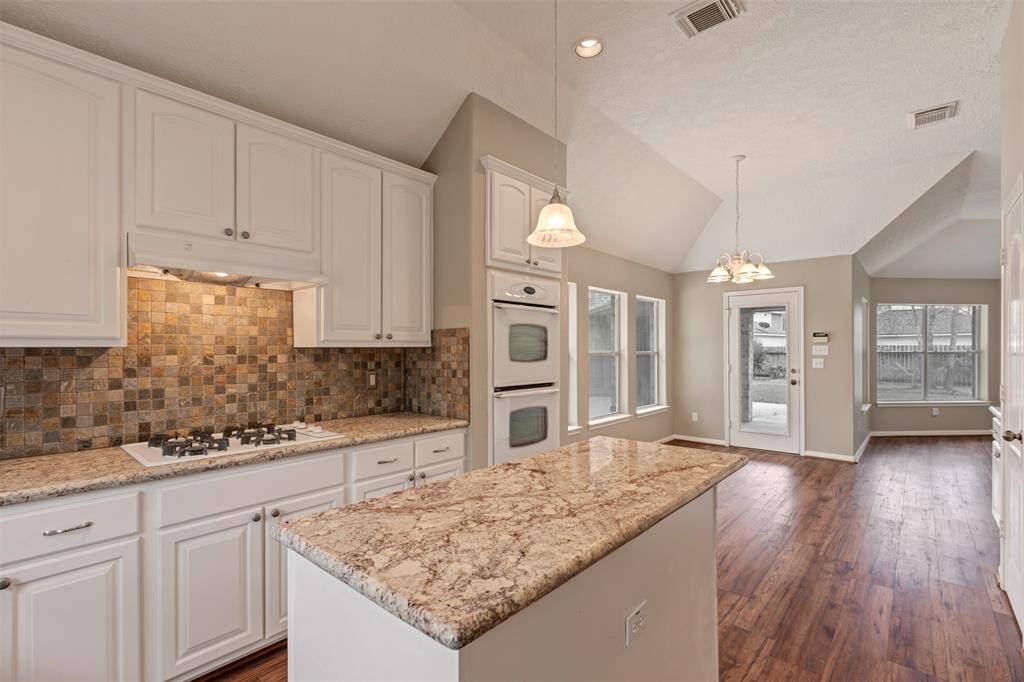 This kitchen features granite countertops, a mosaic tile backsplash, and white cabinetry. It includes a built-in oven and cooktop, with ample natural light from large windows. The open layout flows into a dining area with hardwood flooring and a view of the backyard through glass doors.