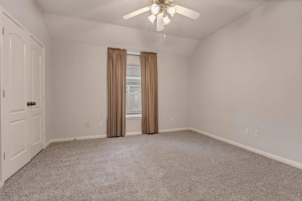 This room features neutral gray walls and carpet, a ceiling fan with lights, a window with beige curtains, and a double-door closet, creating a cozy and versatile space.