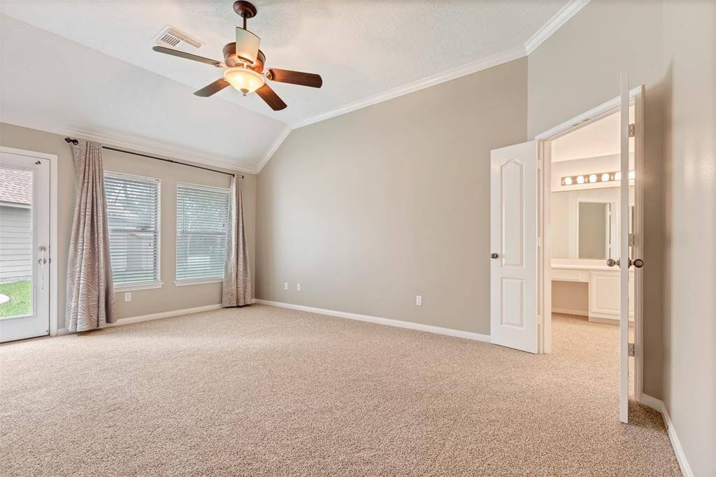 This spacious bedroom features neutral carpet and walls, a ceiling fan, and ample natural light from large windows and a glass door leading outside. It connects to a well-lit bathroom with a vanity.