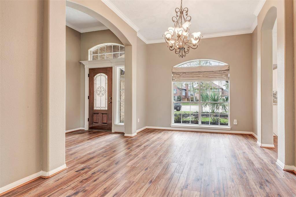 This inviting home entrance features a warm, open space with elegant hardwood flooring, high ceilings, and a beautiful chandelier. Large windows provide ample natural light, and the wooden front door adds a touch of classic charm.