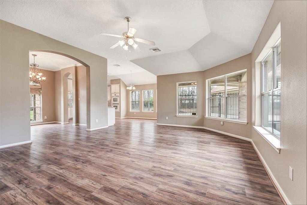 This spacious, open-concept living area features elegant wood flooring, abundant natural light from large windows, and modern ceiling fans. The adjoining dining area has stylish chandeliers and archways, leading into a well-lit kitchen, creating a warm and inviting atmosphere perfect for entertaining.