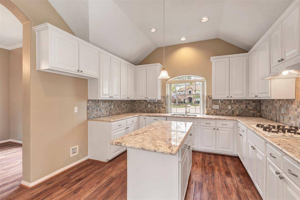 This kitchen features high vaulted ceilings, abundant white cabinetry, and granite countertops. The space includes a central island, a modern gas cooktop, and a large window providing ample natural light. The wooden flooring adds warmth and elegance.