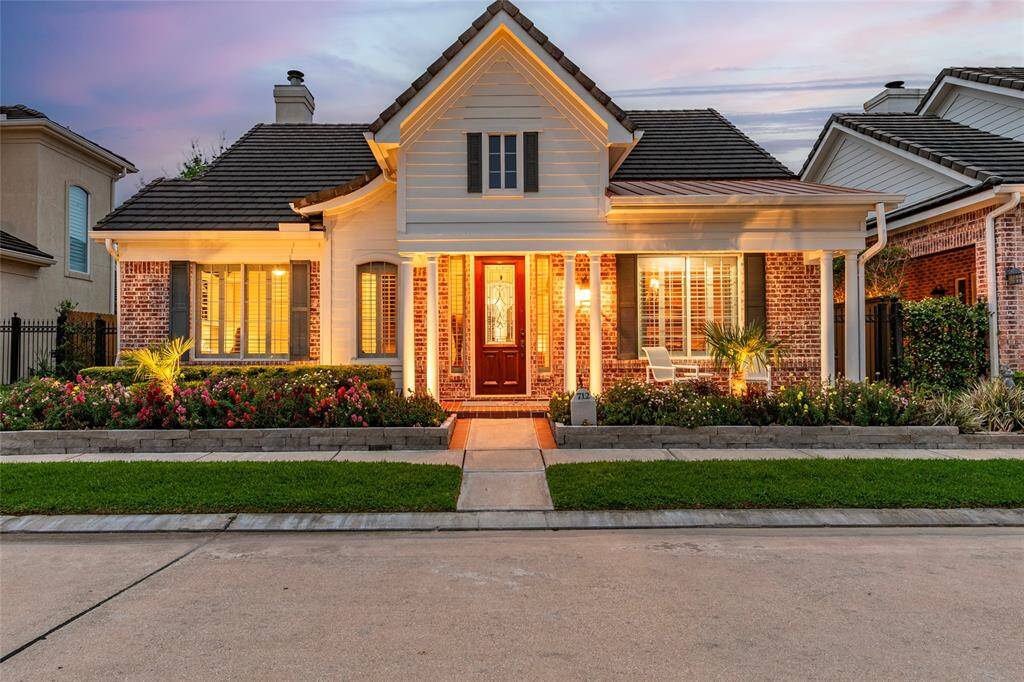 Custom home features a welcoming front porch, brick exterior, and a well-maintained landscaping. The warm lighting and manicured lawn enhance the curb appeal.