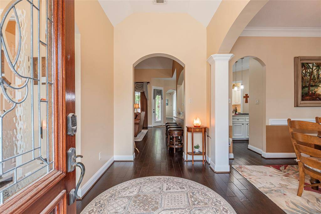 This inviting entryway features a wooden door with glass accents, leading into a bright space with high ceilings and elegant archways. Rich wood flooring runs throughout, connecting to a cozy dining area and a glimpse of the living room beyond.