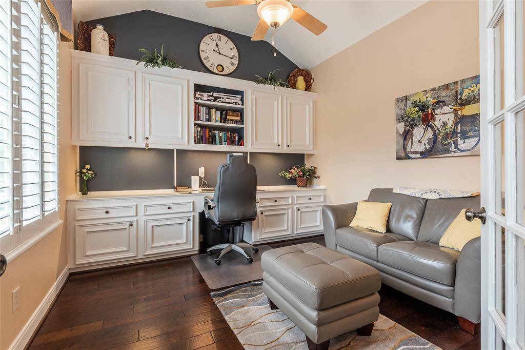 This cozy home office features built-in white cabinets, a desk with shelving, and a comfortable seating area with a leather sofa and ottoman. Natural light streams in through plantation shutters, highlighting the warm wood flooring and high ceilngs.