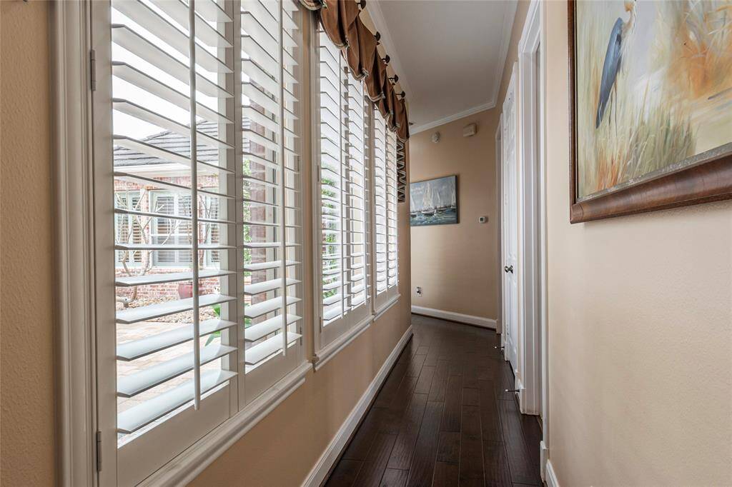 Bright hallway with large windows featuring white plantation shutters, offering views of a courtyard. The space has wood flooring and neutral walls adorned with artwork, creating an inviting atmosphere.