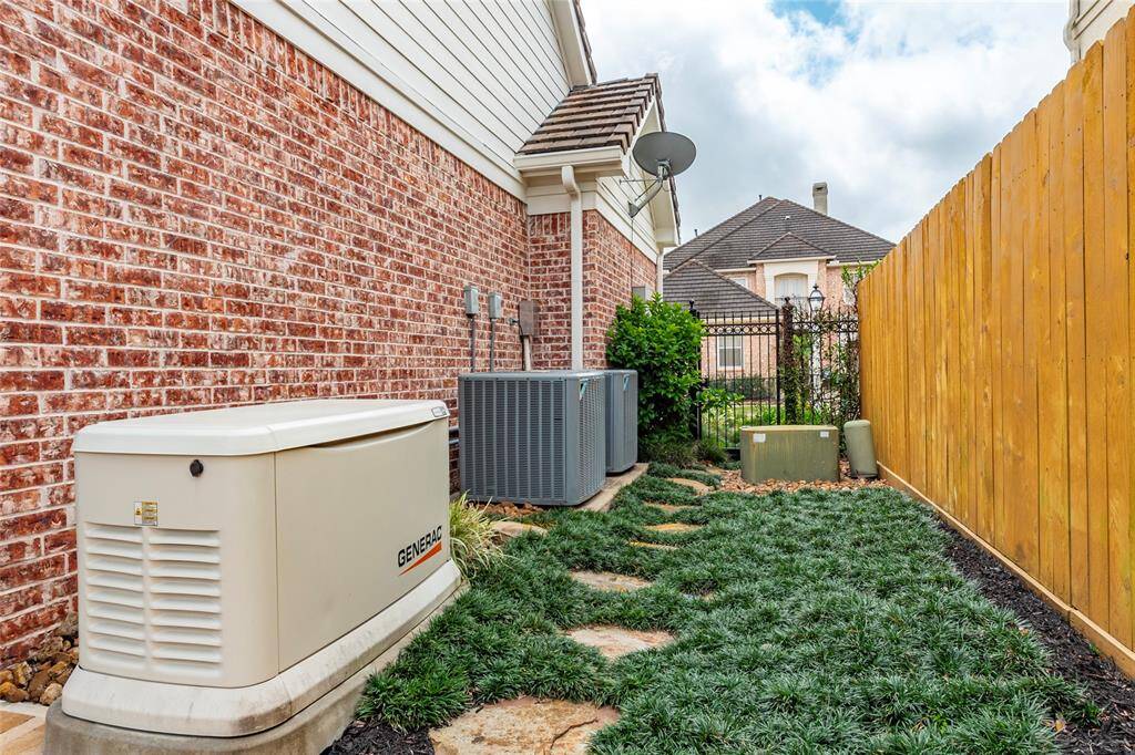 This photo shows a side yard with a brick exterior wall, featuring a backup generator and updated HVAC units. The area is landscaped with stepping stones and a wooden fence for privacy.