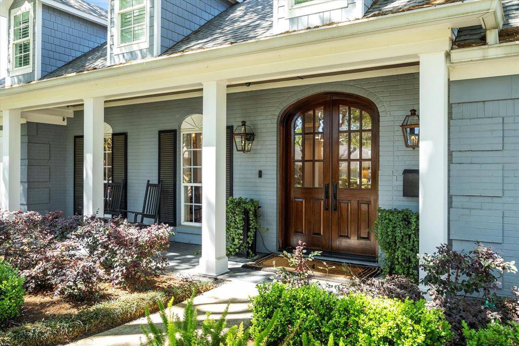 Charming home with inviting front porch, featuring elegant wooden double doors and classic lantern lighting. The exterior is accented with tasteful landscaping and a cozy rocking chair area, perfect for relaxation.