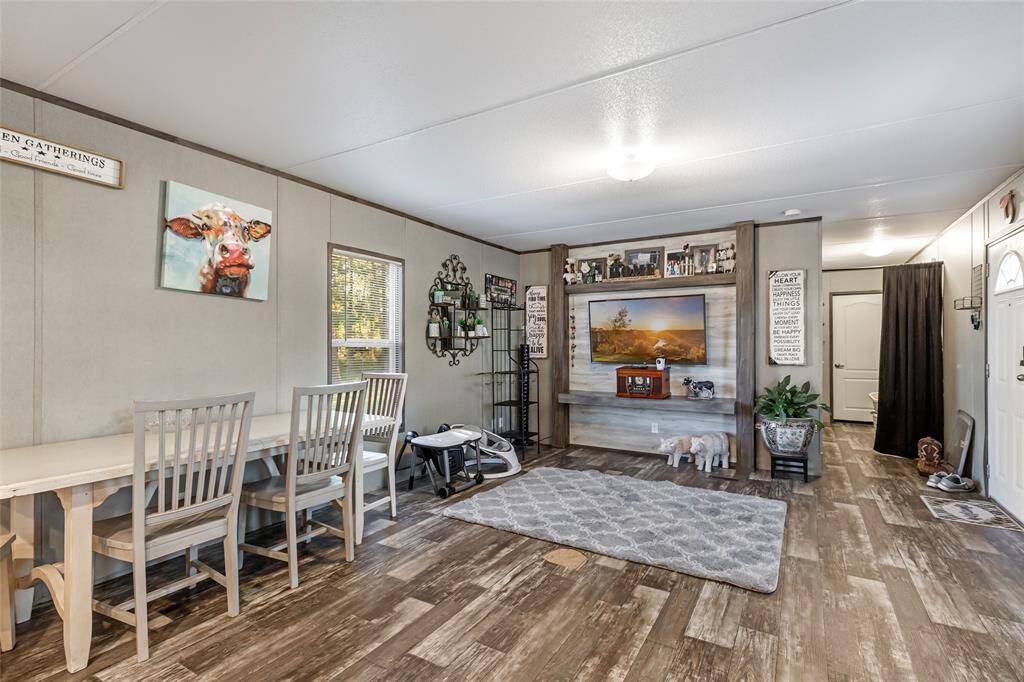 This cozy living space features a rustic design with wood-look flooring and a neutral color palette. It includes a dining area with a farmhouse-style table, a comfortable sitting area with a large TV, and decorative accents like artwork and plants. The room is well-lit with natural light from the window.