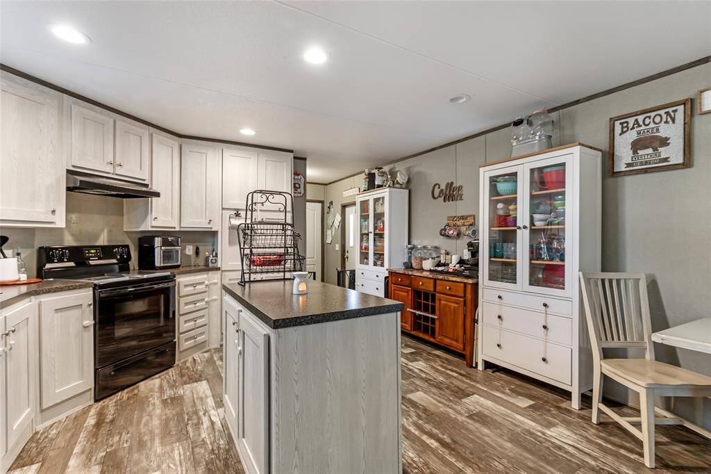 This kitchen features modern white cabinetry, a central island with a dark countertop, and modern appliances. The space is well-lit with recessed lighting, and there is ample storage with glass-front cabinets and additional shelving, creating a cozy and functional cooking area.