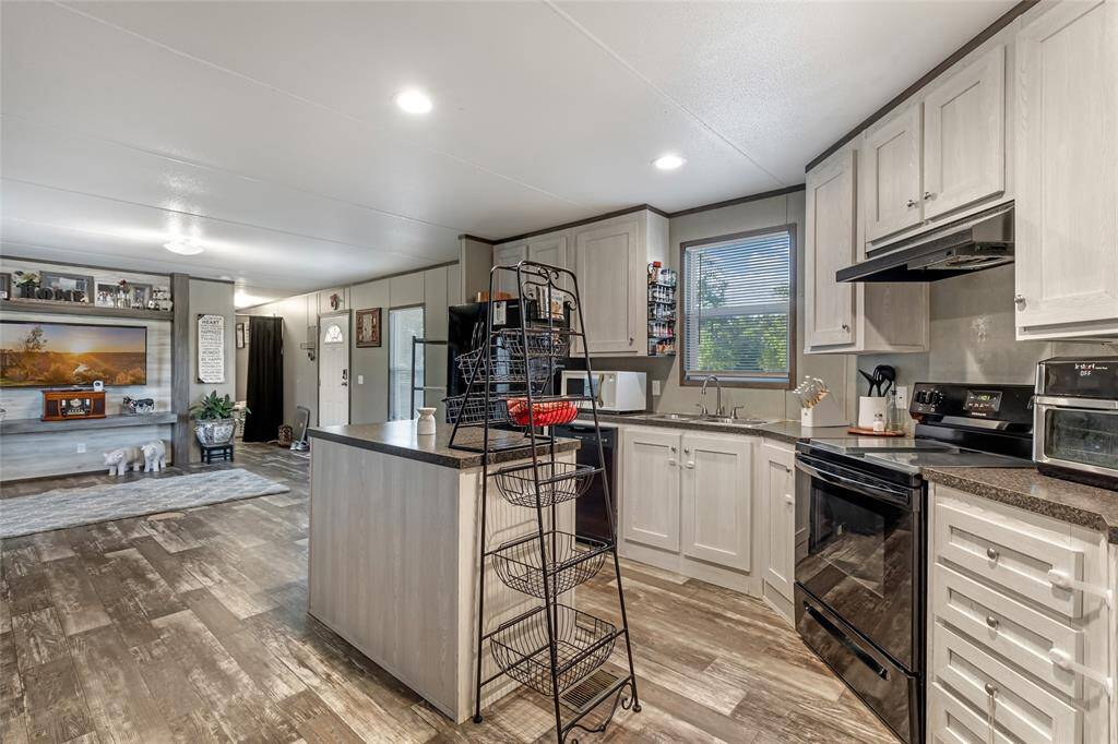 This photo showcases a modern, open-concept kitchen with light cabinetry, dark countertops, and stainless steel appliances. The kitchen opens to a cozy living area with wood-style flooring and ample natural light.