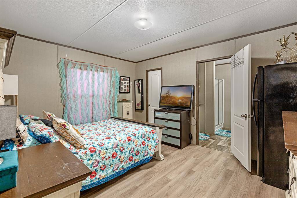 This cozy bedroom features a floral bedspread, light wood flooring, and neutral walls. It includes a TV on a dresser, a window with colorful curtains, and an ensuite bathroom. There's also a fridge, adding convenience to the space.
